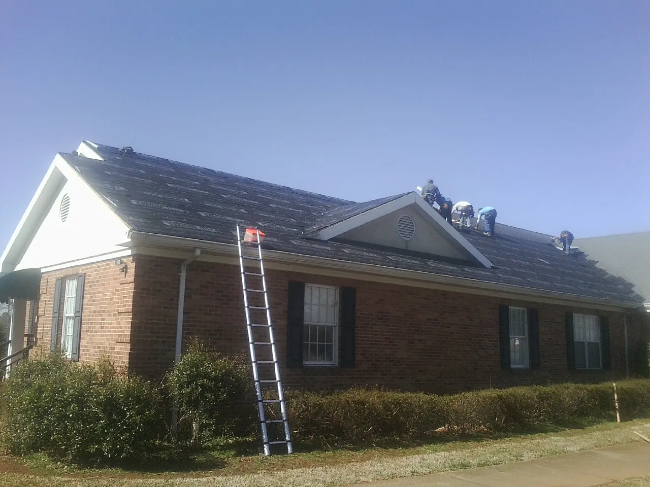 Crew tearing shingles off a brick ranch roof with a ladder set — Emergency Roof Repair work by our crew in Hector