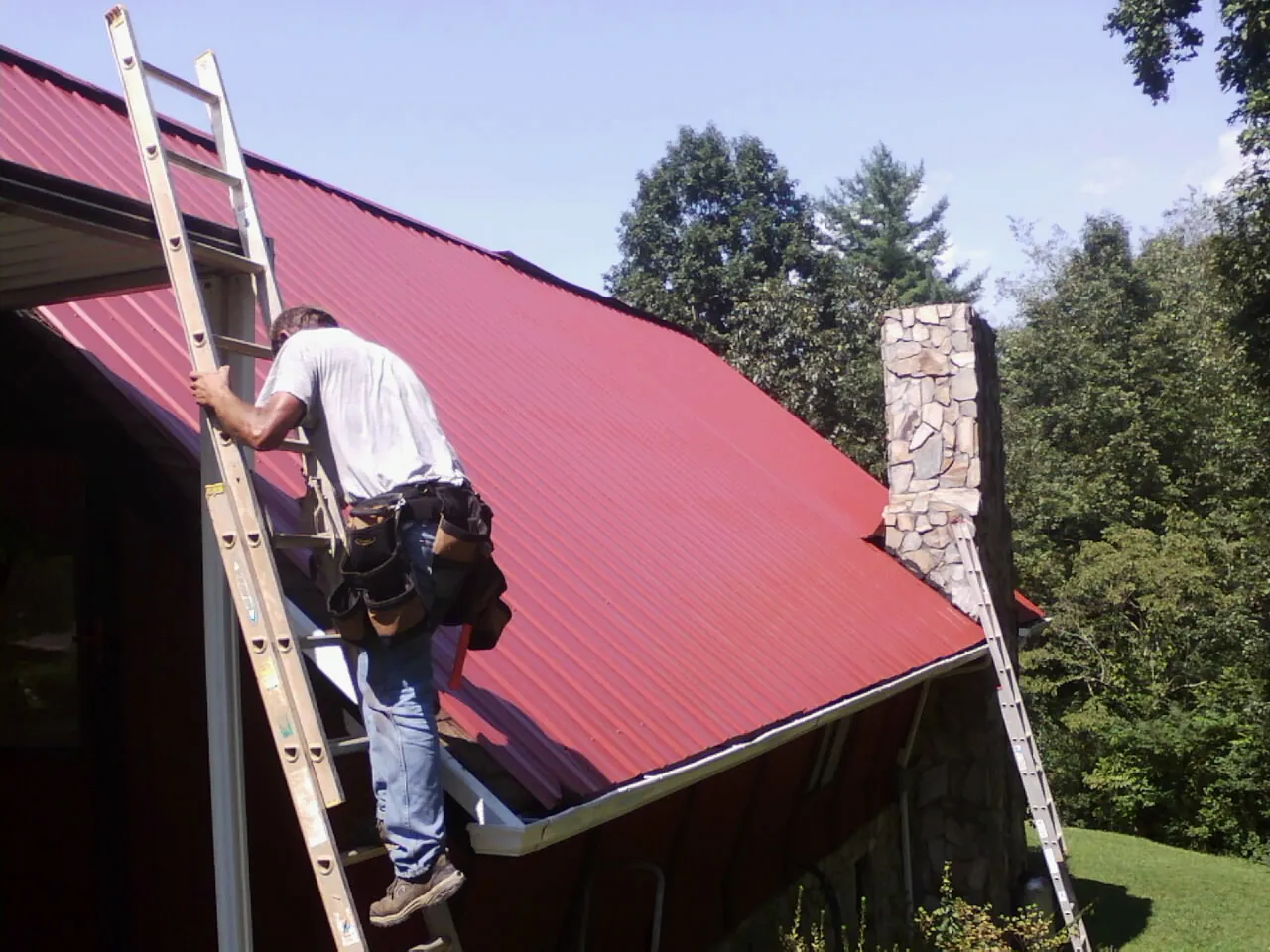 Roofer on a ladder beside a red metal roof and stone chimney — Soffit & Fascia Repair by Roofing Brothers in Hector